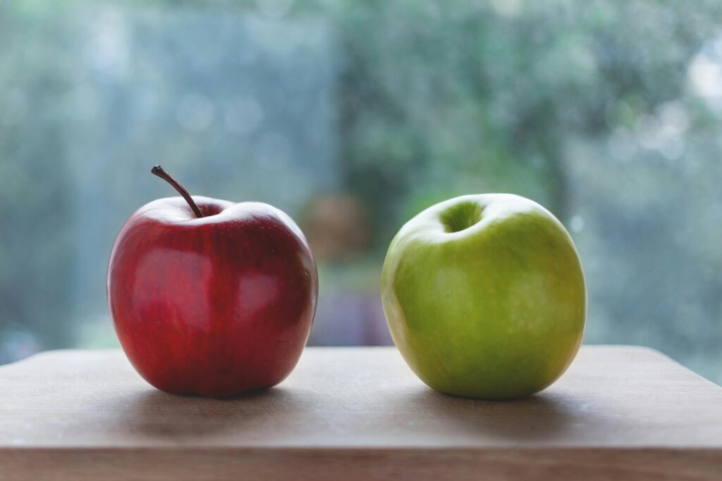 Two fresh apples, one red and one green, sitting on a wooden surface indoors.